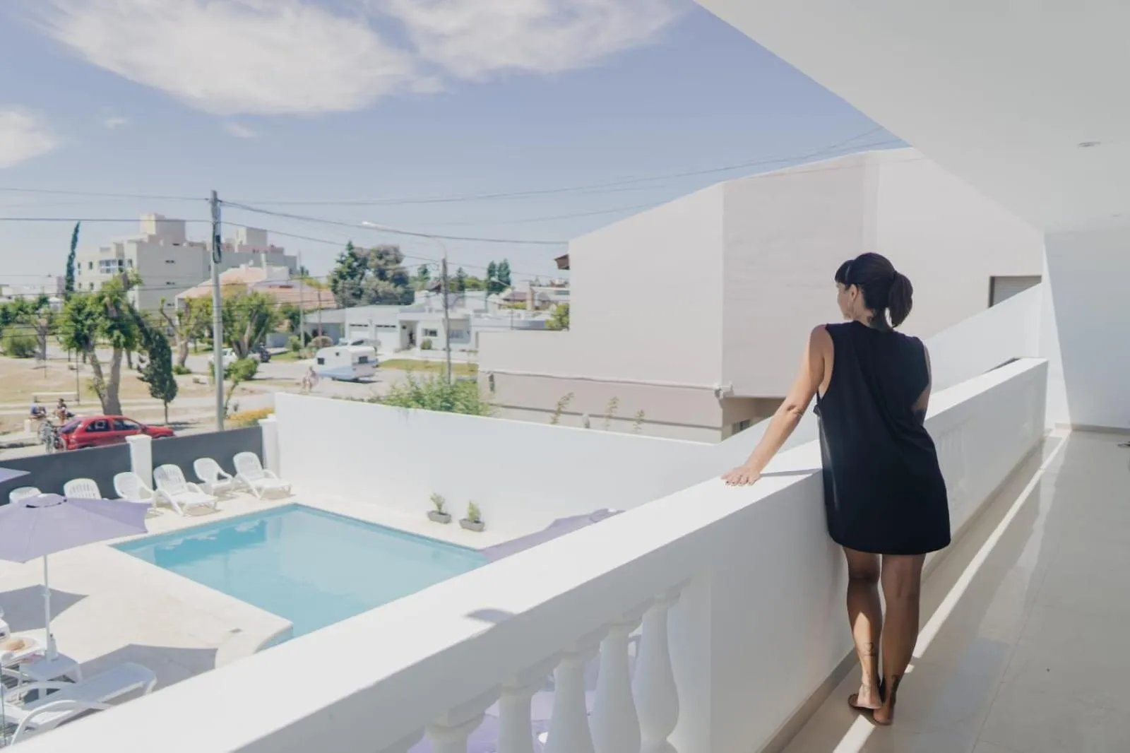 Une femme se tient sur une terrasse blanche avec vue sur une piscine et un paysage urbain.