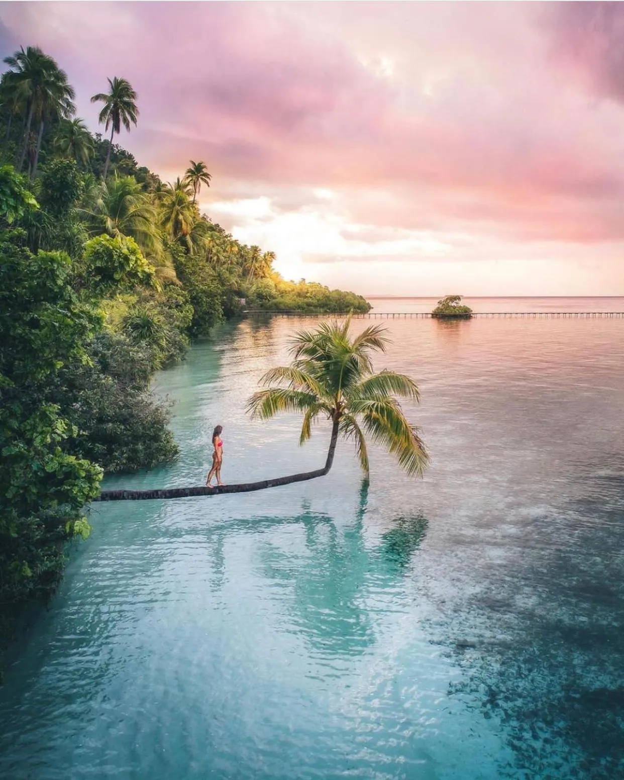 Une personne se tient debout sur une roche au bord de l'eau turquoise, face à un coucher de soleil rose et orangé.