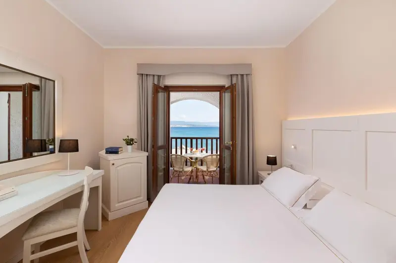 Chambre lumineuse avec un lit blanc et une vue sur la mer depuis un balcon.