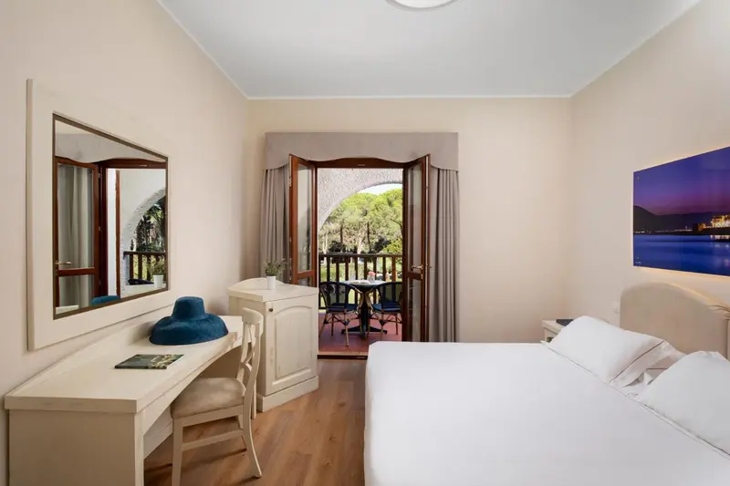 Une chambre lumineuse avec un lit blanc et un bureau près d'une fenêtre ouverte sur une terrasse.
