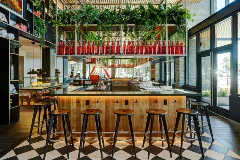 Un bar lumineux avec un comptoir en bois et des tabourets, décoré de plantes suspendues et de bouteilles colorées.