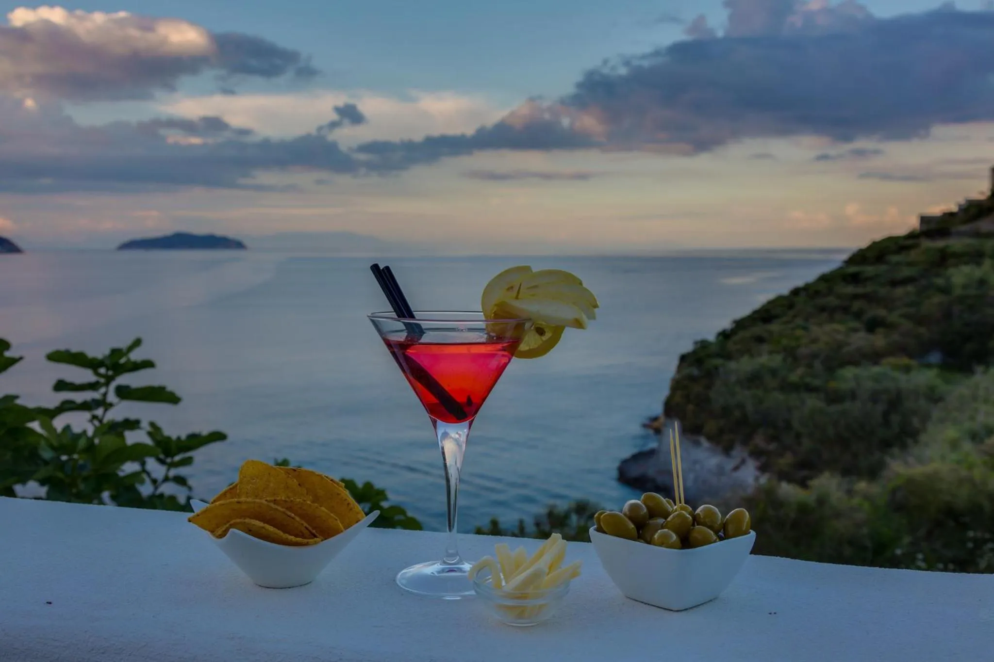 Un cocktail rouge vif et des amuse-gueules sont servis sur une terrasse avec vue sur la mer.