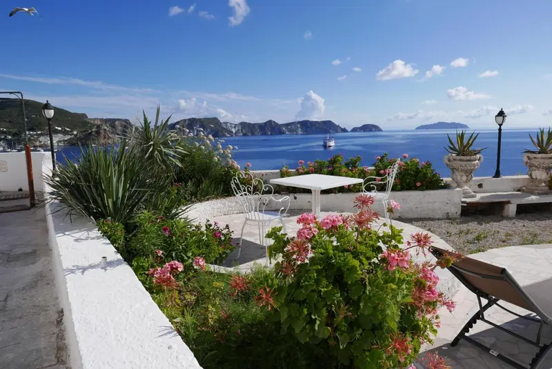 Terrasse fleurie avec mobilier blanc offrant une vue sur la mer et les collines.