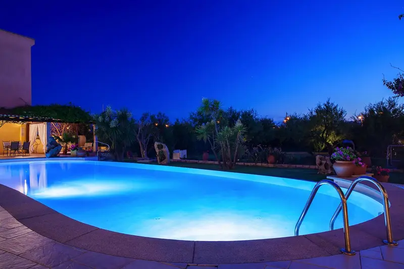 Piscine illuminée au crépuscule avec végétation luxuriante et éclairage d'ambiance.