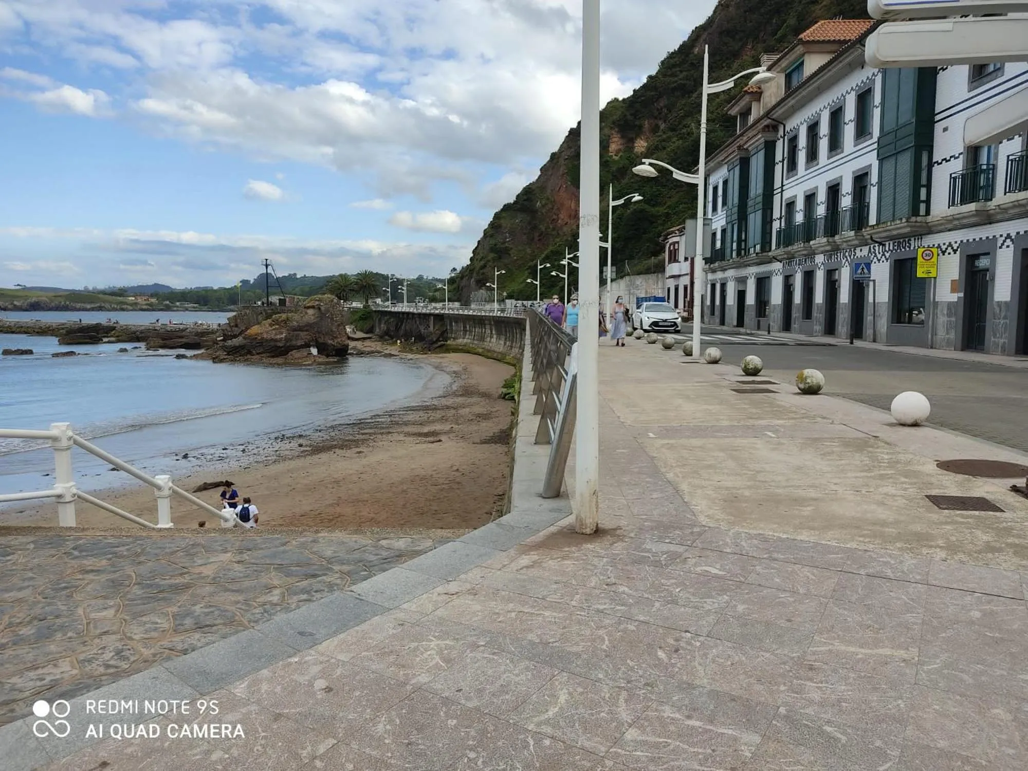 Vue d'une promenade en bord de mer avec une plage de sable et des bâtiments colorés sur une colline.