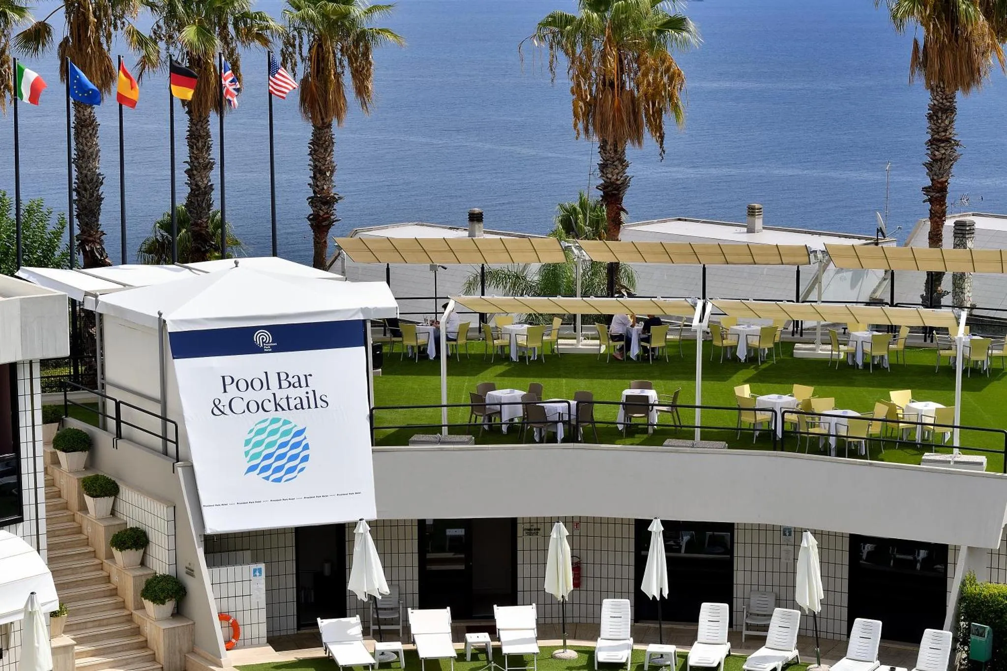 Terrasse avec tables et parasols donnant sur la mer et des drapeaux.