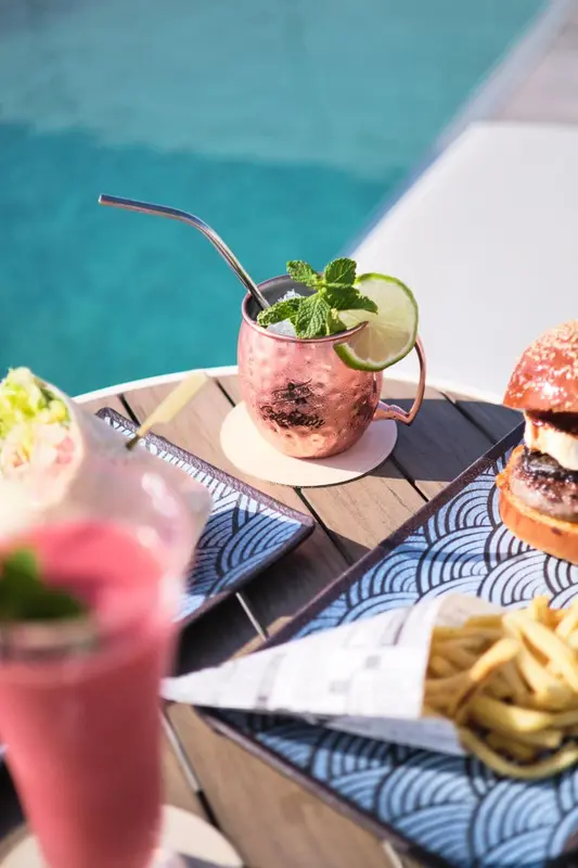 Un cocktail et un repas sont servis sur une table en bois près d'une piscine.