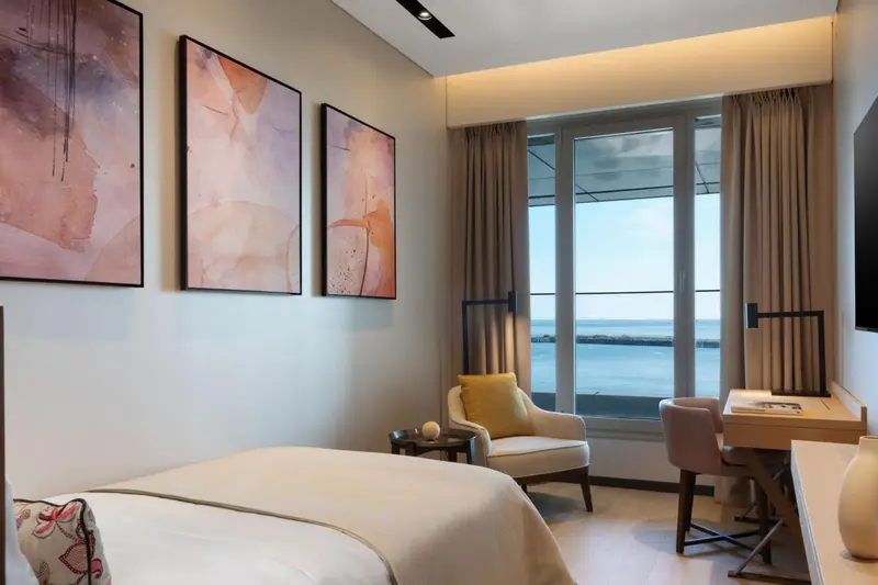 Une chambre lumineuse avec un lit, un fauteuil et un bureau donnant sur une vue mer.