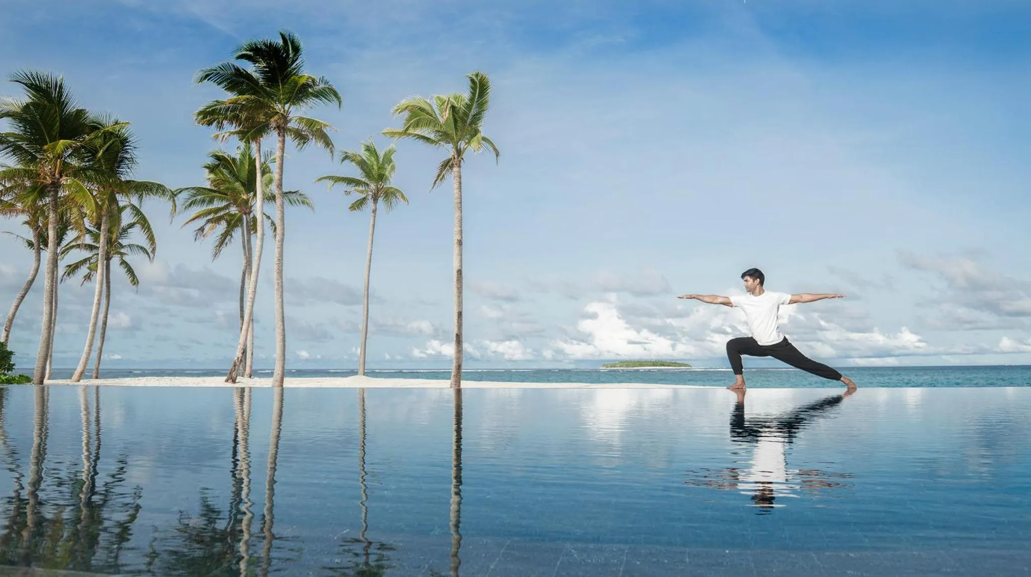 Une personne pratique le yoga au bord d'une piscine à débordement avec une vue sur la mer et des palmiers.