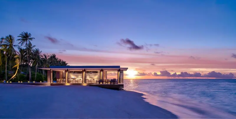 Un restaurant sur pilotis s'ouvre sur l'océan au coucher du soleil.