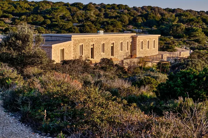 Bâtiment bas en pierre avec une terrasse en contrebas, entouré de végétation.