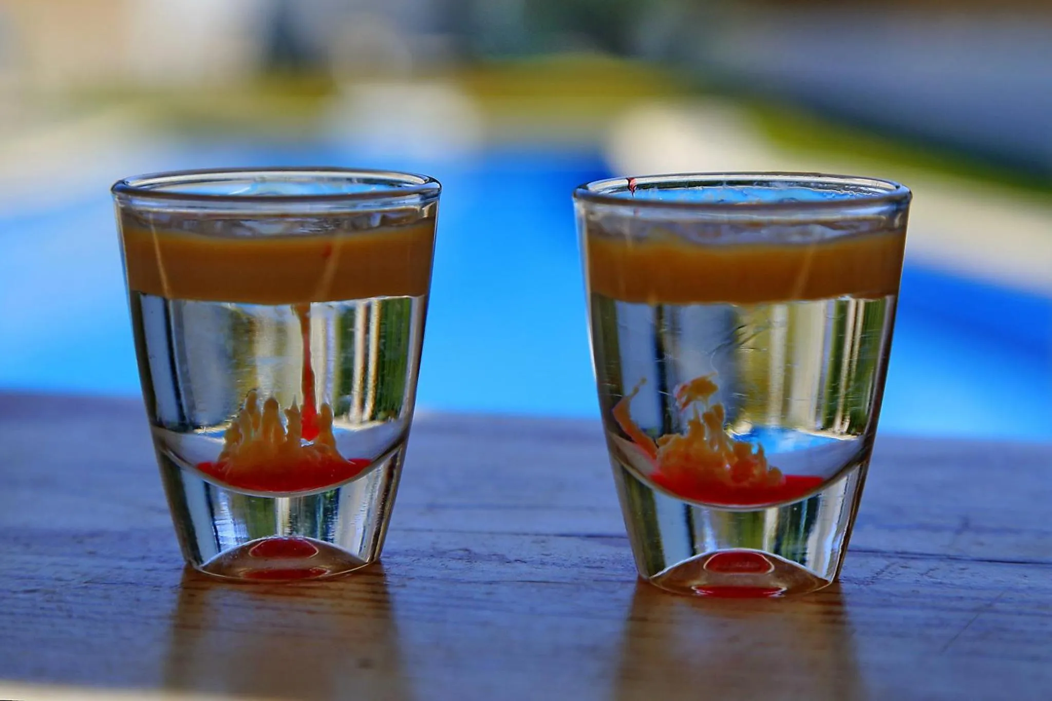 Deux verres à shooter colorés sont posés sur une table en bois devant une piscine.