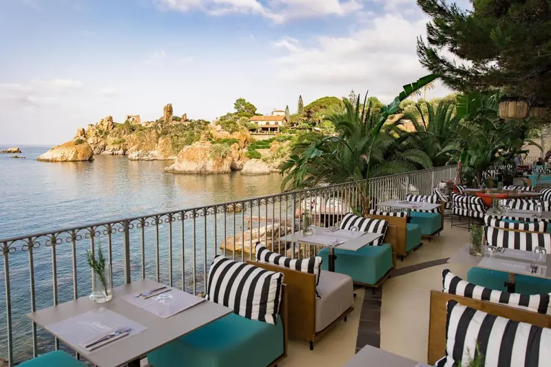 Tables et chaises sur une terrasse ensoleillée avec vue sur la mer et les rochers.