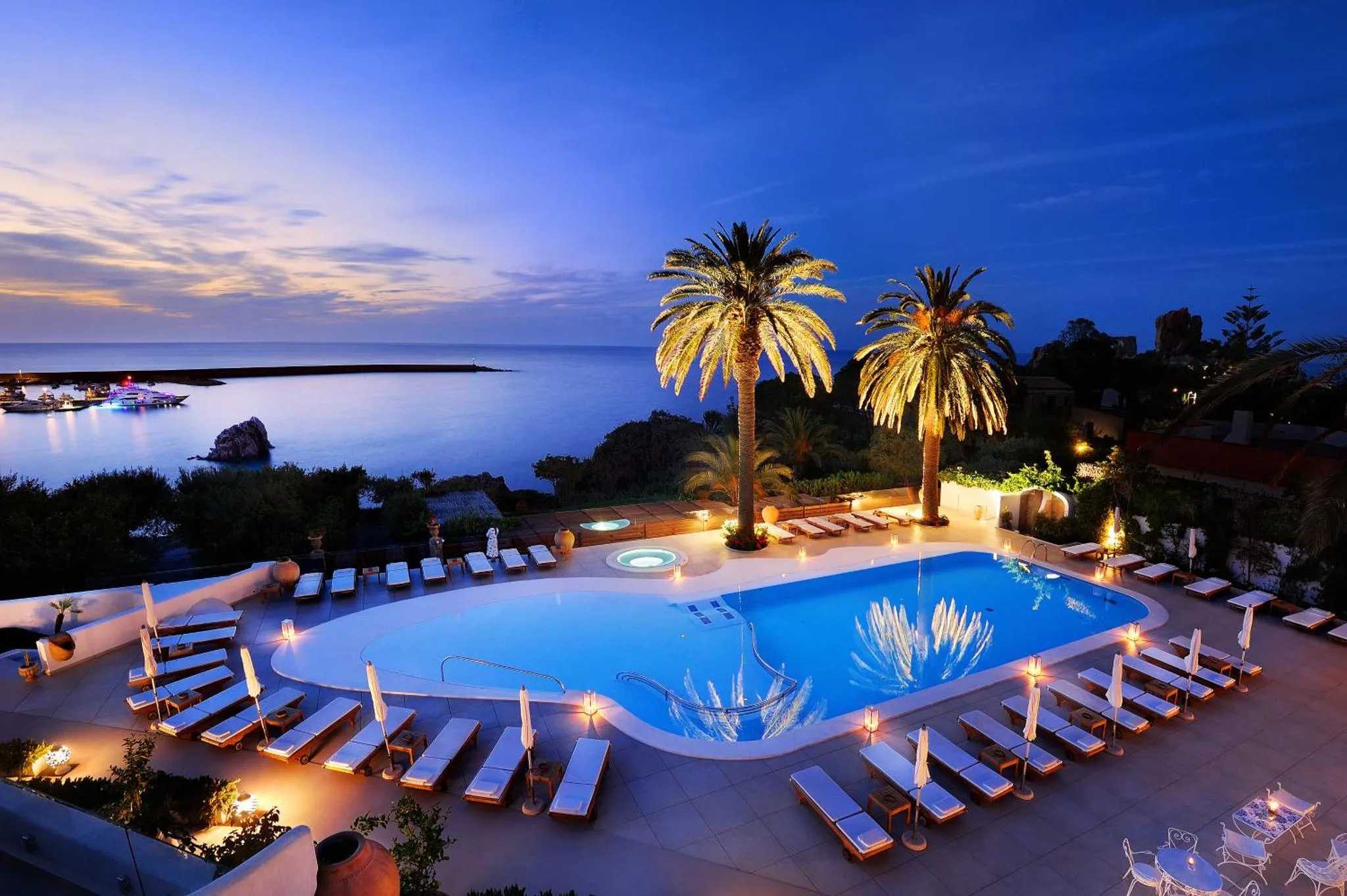 Piscine illuminée avec transats et palmiers, vue sur la mer au crépuscule.