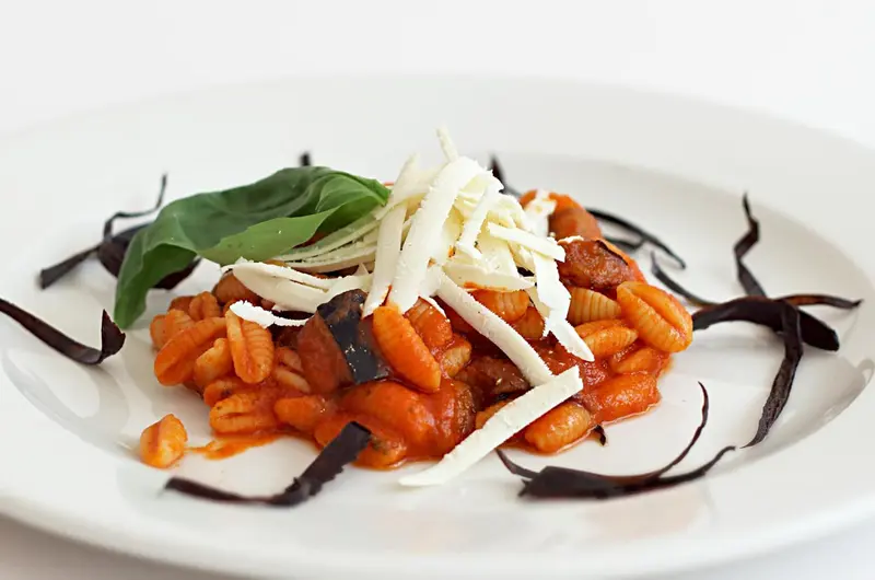 Assiette de pâtes aux aubergines et à la sauce tomate, décorée de basilic frais et de copeaux de fromage.