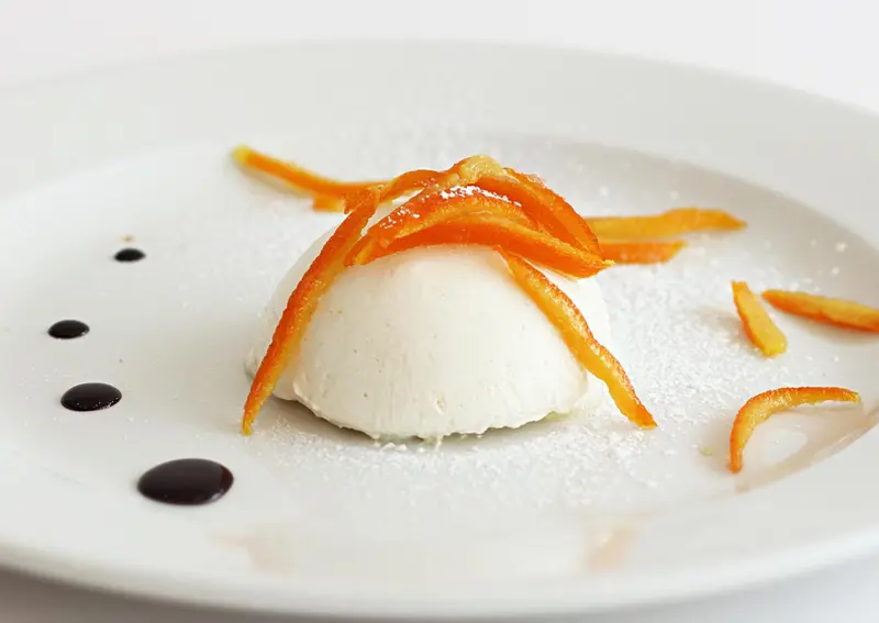 Une portion de dessert blanc est décorée de zestes d'orange et d'une sauce foncée.