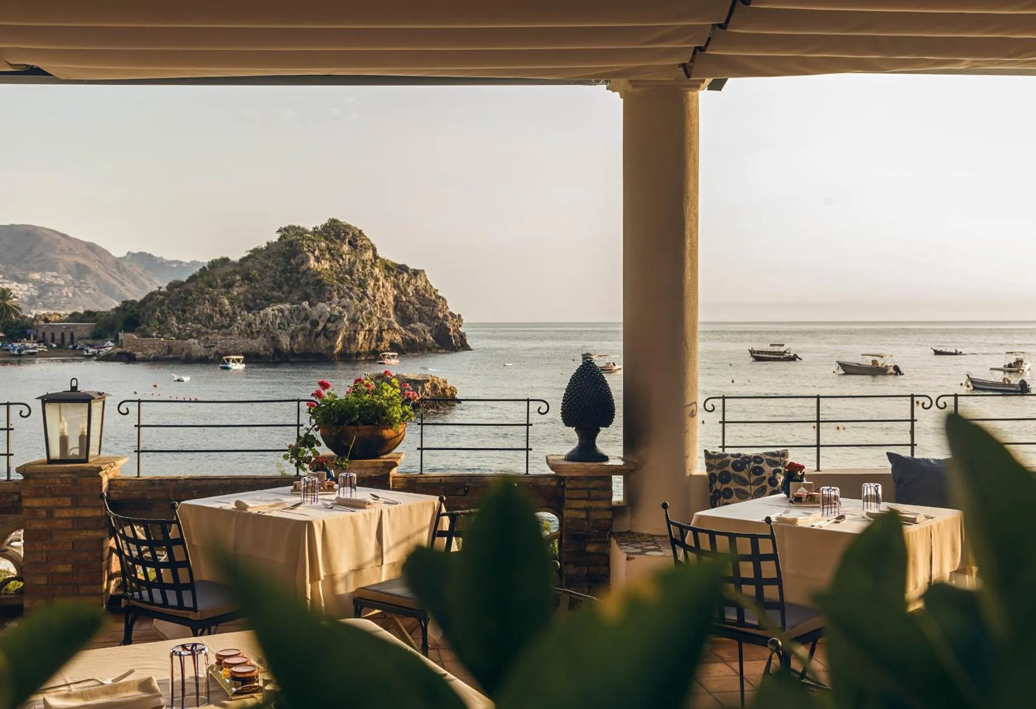 Terrasse ombragée avec tables nappées donnant sur la mer et une côte rocheuse.