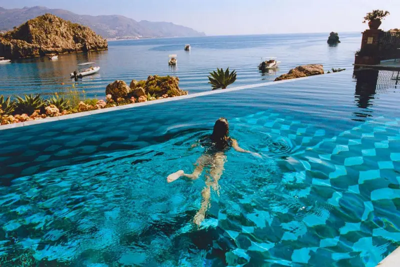 Une personne nage dans une piscine à débordement avec vue sur la mer et des formations rocheuses.