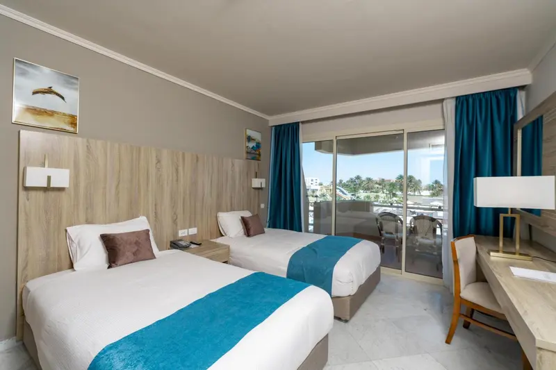 Chambre lumineuse avec deux lits et vue sur la mer depuis un balcon.