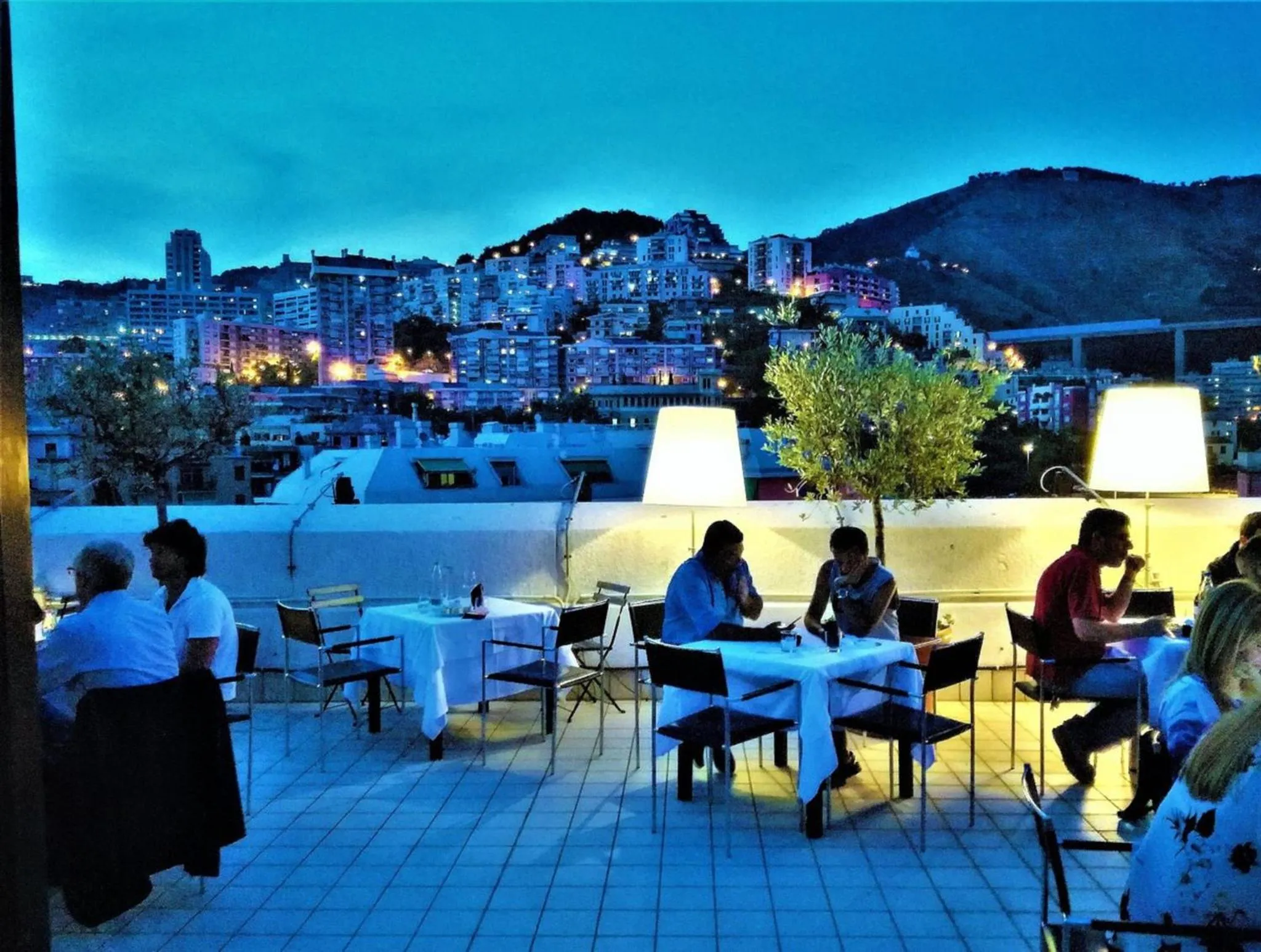Terrasse lumineuse avec des tables et des convives face à une ville illuminée.