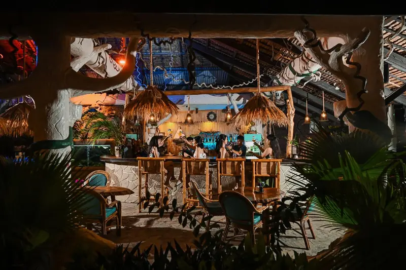 Un comptoir de bar éclairé accueille des clients dans un espace extérieur tropical.