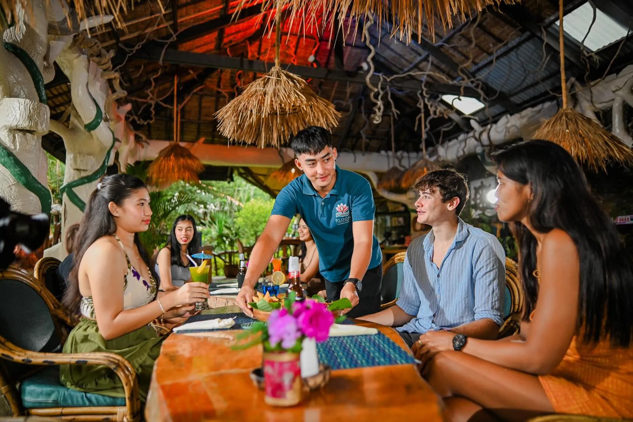 Des clients sont servis à une table en bois dans un espace ouvert décoré de chaume.