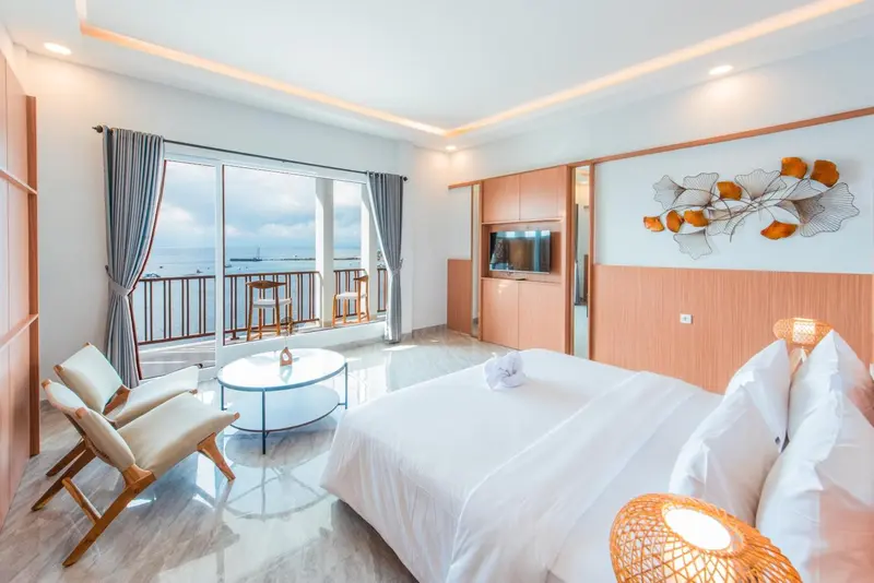 Chambre lumineuse avec un lit blanc et une vue sur la mer par de grandes baies vitrées.