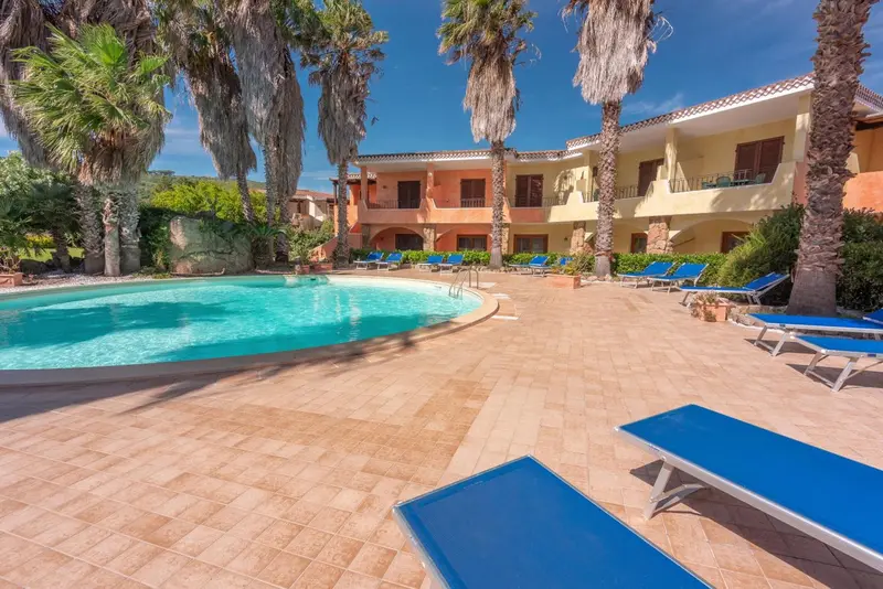 Bâtiment à deux étages avec piscine et transats sous des palmiers.