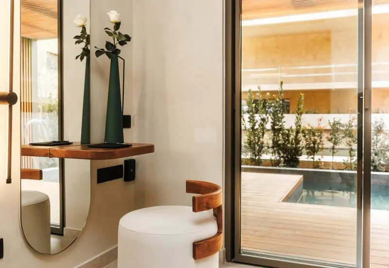 Une coiffeuse blanche et un tabouret clair se trouvent près d'une grande baie vitrée donnant sur une terrasse en bois et une piscine.