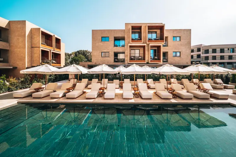 Transats blancs disposés autour d'une piscine turquoise devant un bâtiment beige.