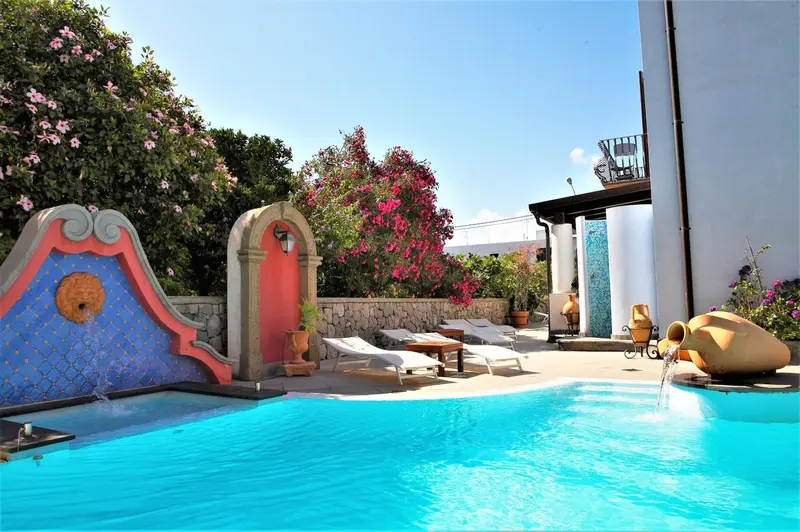 Une piscine turquoise avec fontaine et chaises longues dans un jardin fleuri.