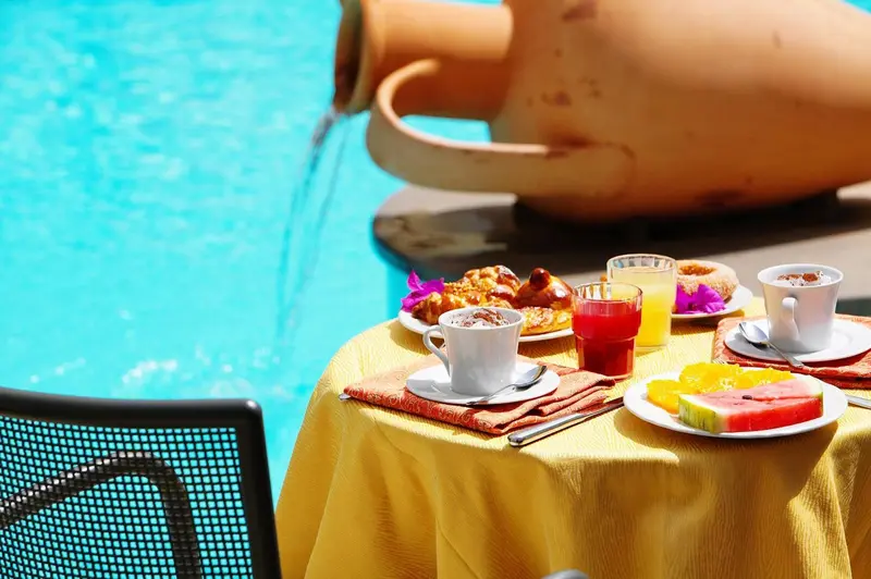 Une table jaune est dressée au bord d'une piscine avec des viennoiseries, des fruits et des boissons.
