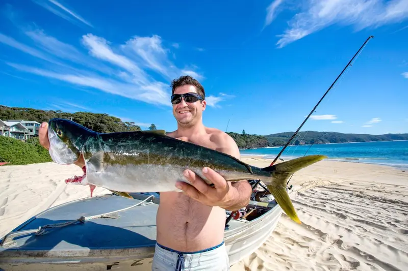 Un homme torse nu tient un poisson qu'il vient de pêcher sur une plage ensoleillée.