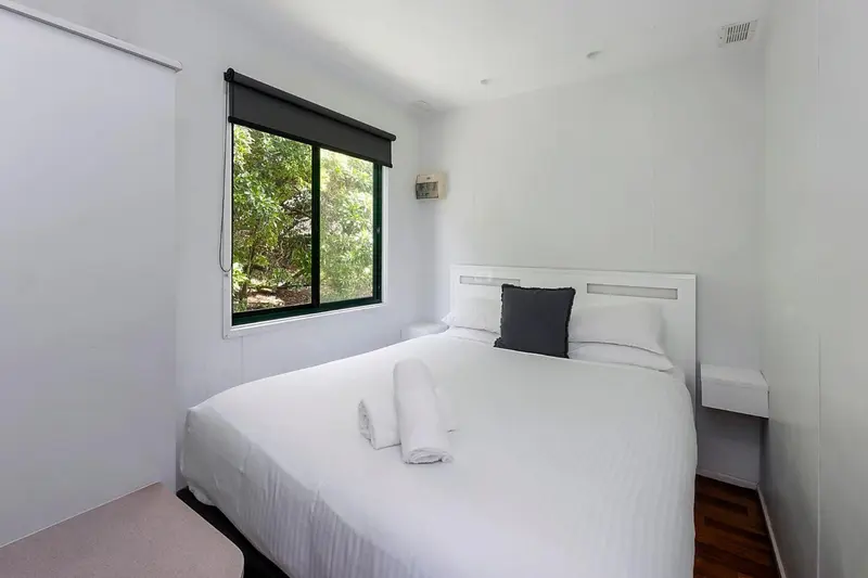 Une chambre lumineuse avec un grand lit blanc et une fenêtre donnant sur la végétation.