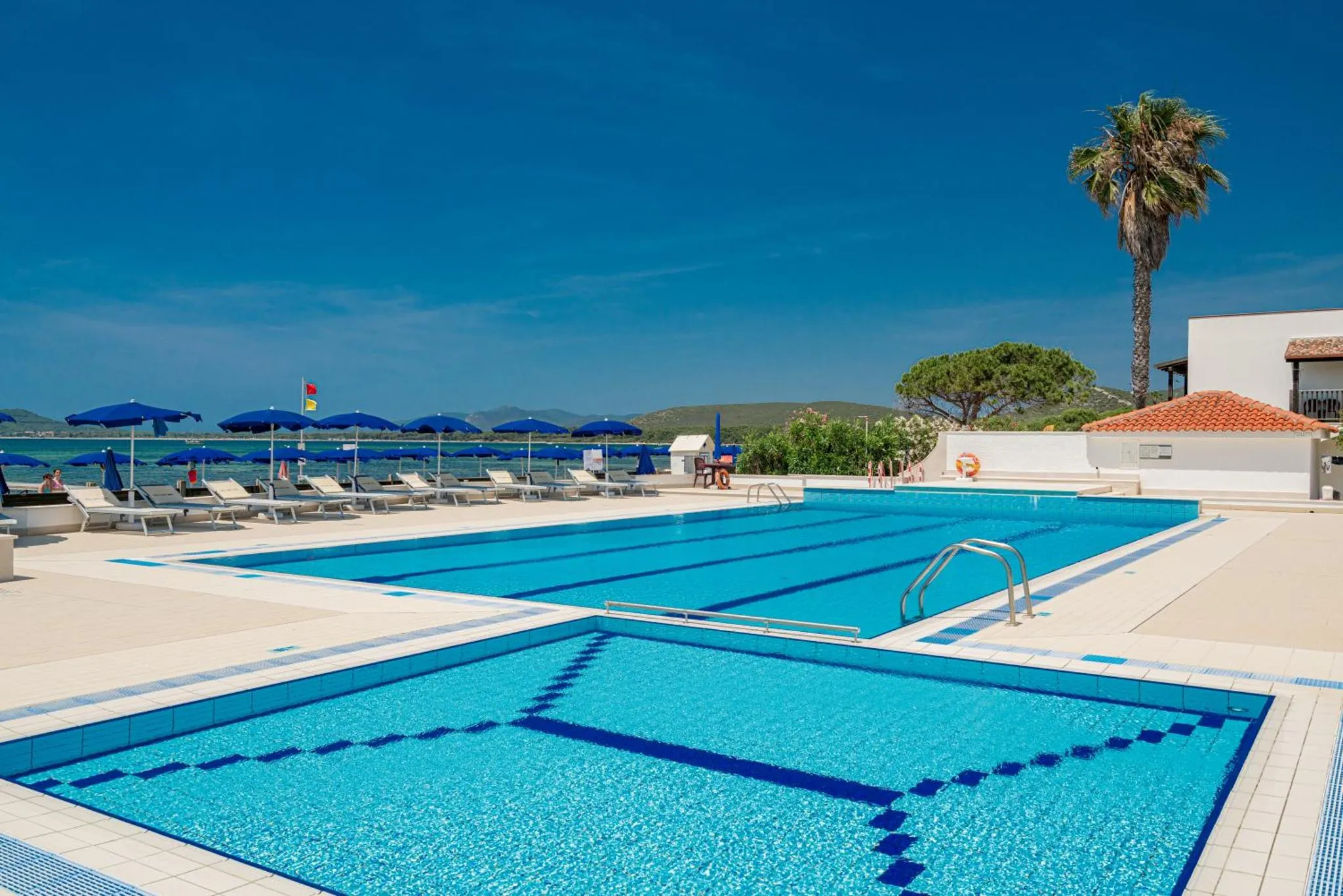 Une piscine rectangulaire turquoise avec des transats bleus et parasols blancs en bord de mer.