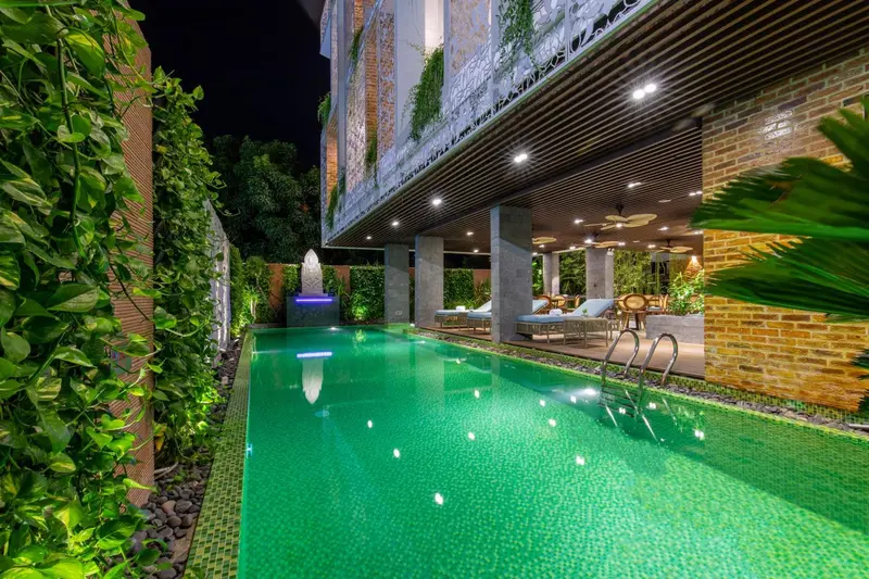 Piscine rectangulaire illuminée avec transats et végétation luxuriante.
