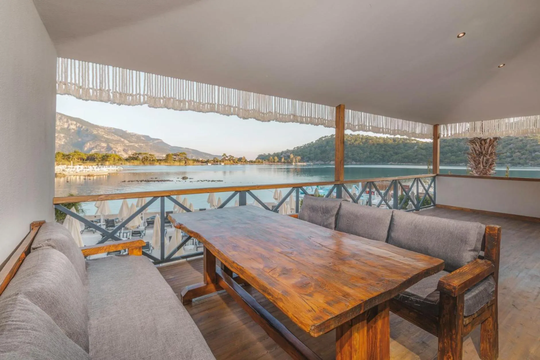 Une table et des banquettes en bois se trouvent sur une terrasse ombragée avec vue sur la mer et les montagnes.
