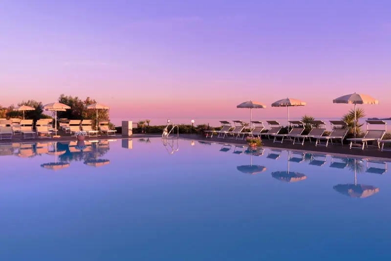 Transats et parasols bordent une piscine reflétant un ciel rosé au coucher du soleil.