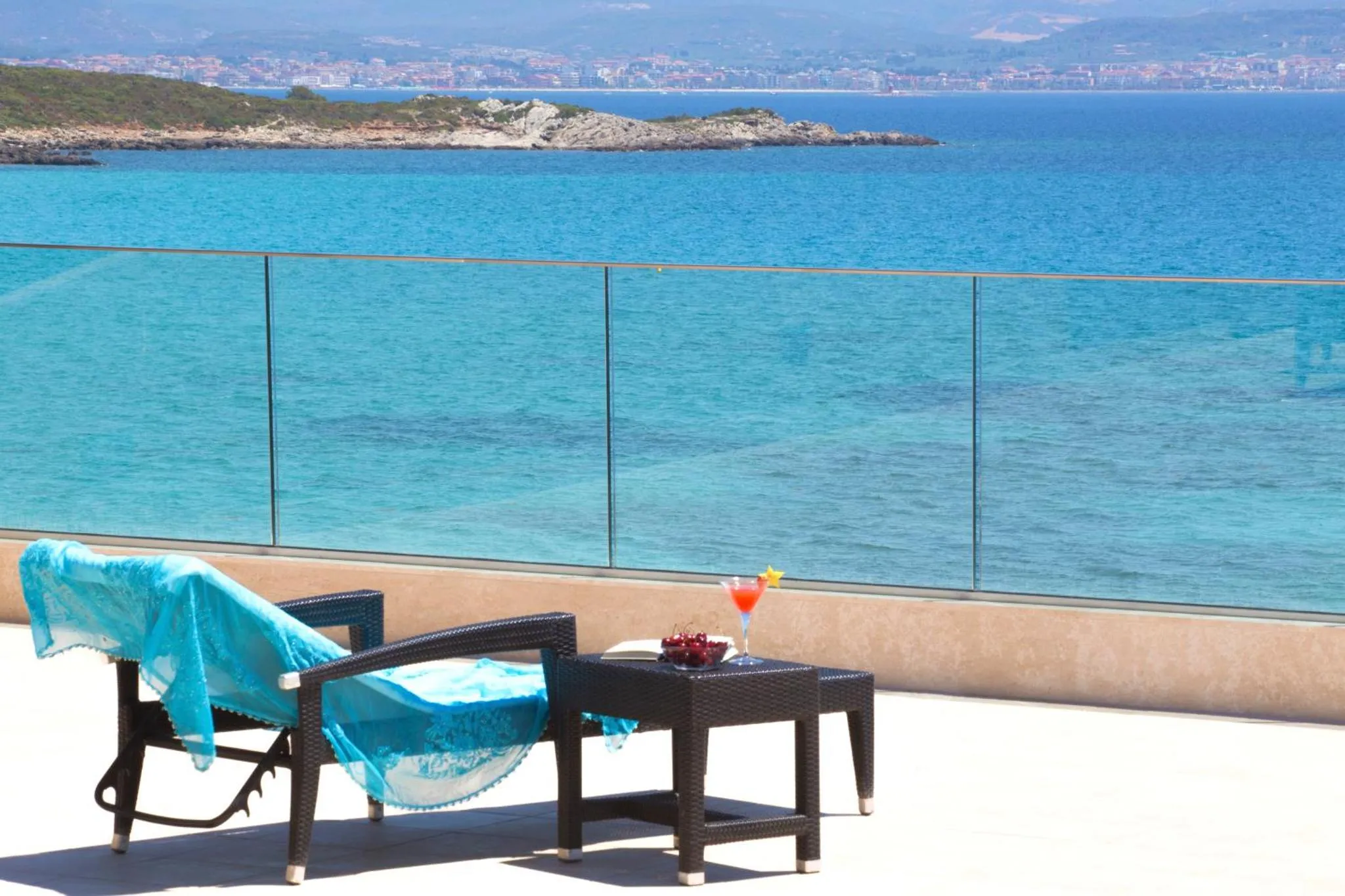 Une chaise longue et une table basse avec des boissons sont posées sur une terrasse ensoleillée avec vue sur la mer.