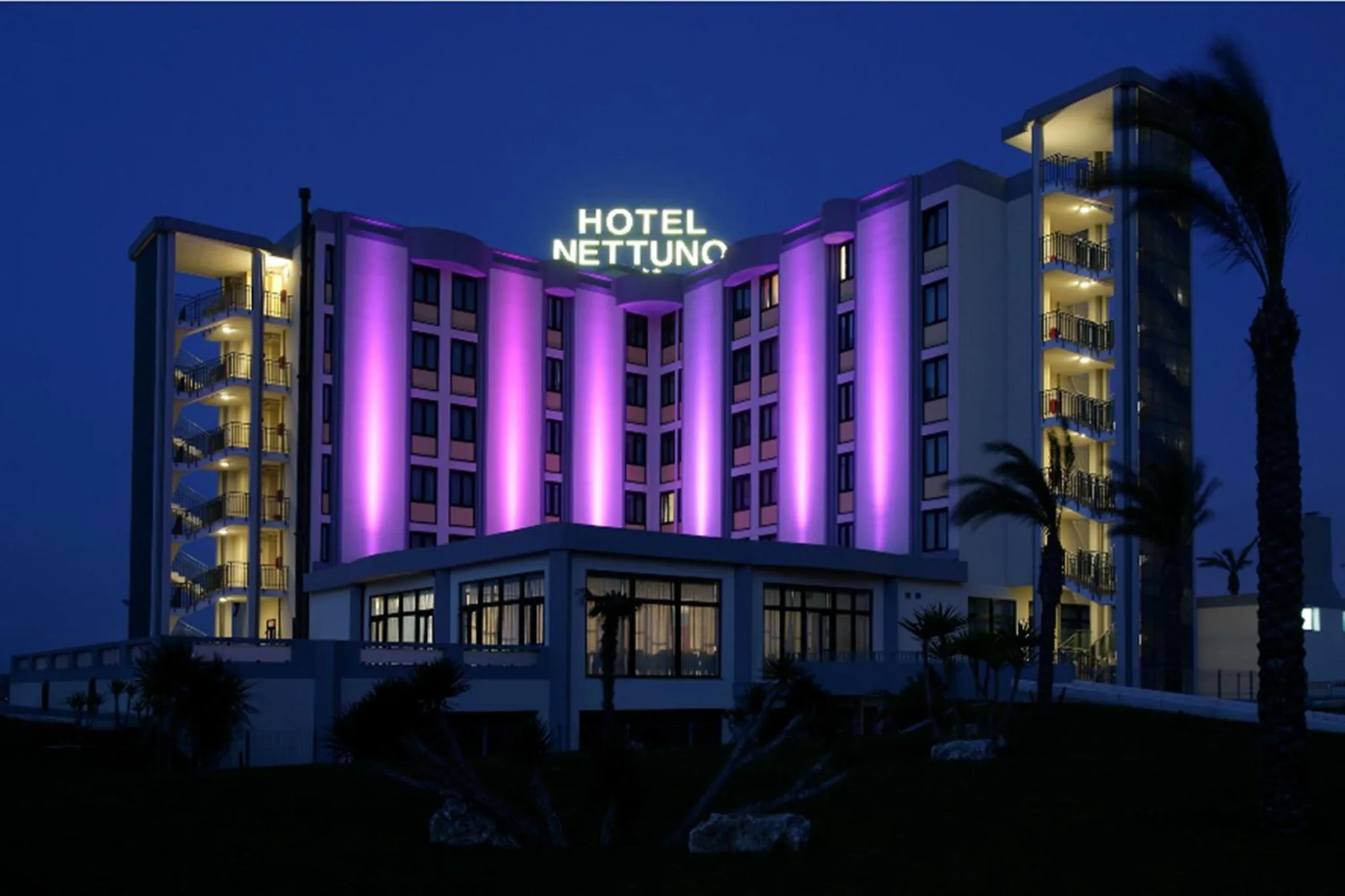 Bâtiment moderne illuminé de couleurs vives au crépuscule, avec des palmiers en premier plan.