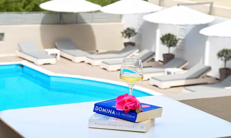 Une piscine turquoise bordée de chaises longues blanches, avec un livre et un verre sur une table.