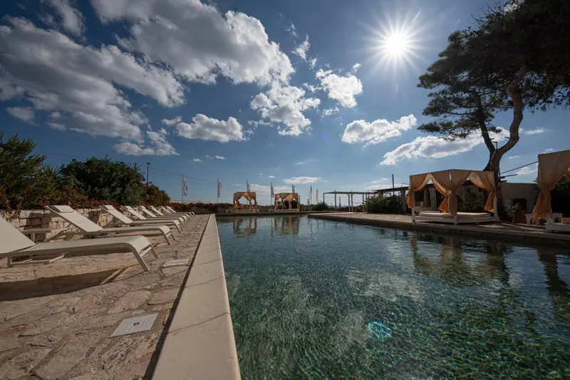 Transats blancs alignés au bord d'une piscine rectangulaire sous un ciel bleu nuageux.