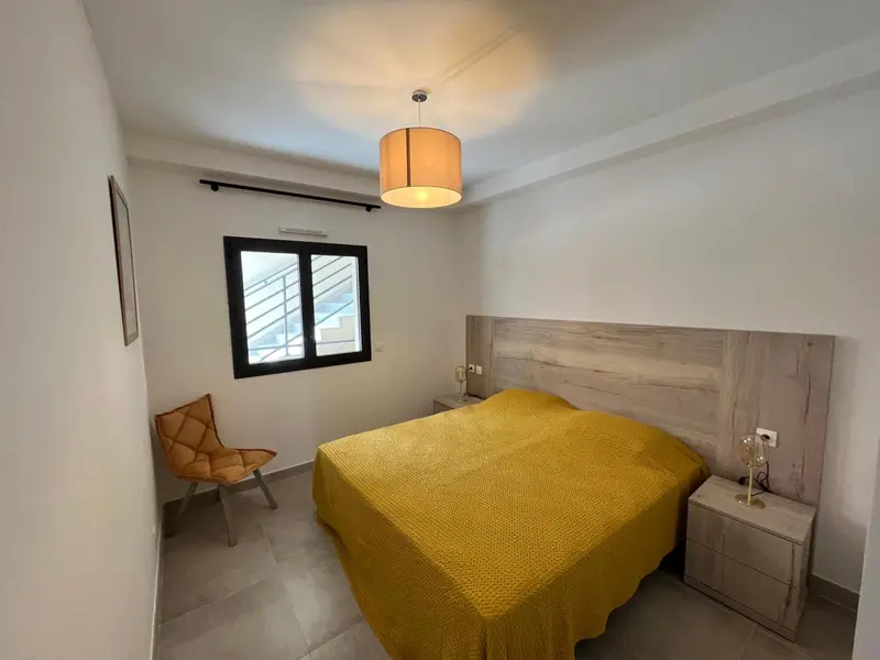 Chambre lumineuse avec un grand lit recouvert d'une couverture jaune et une chaise en osier.