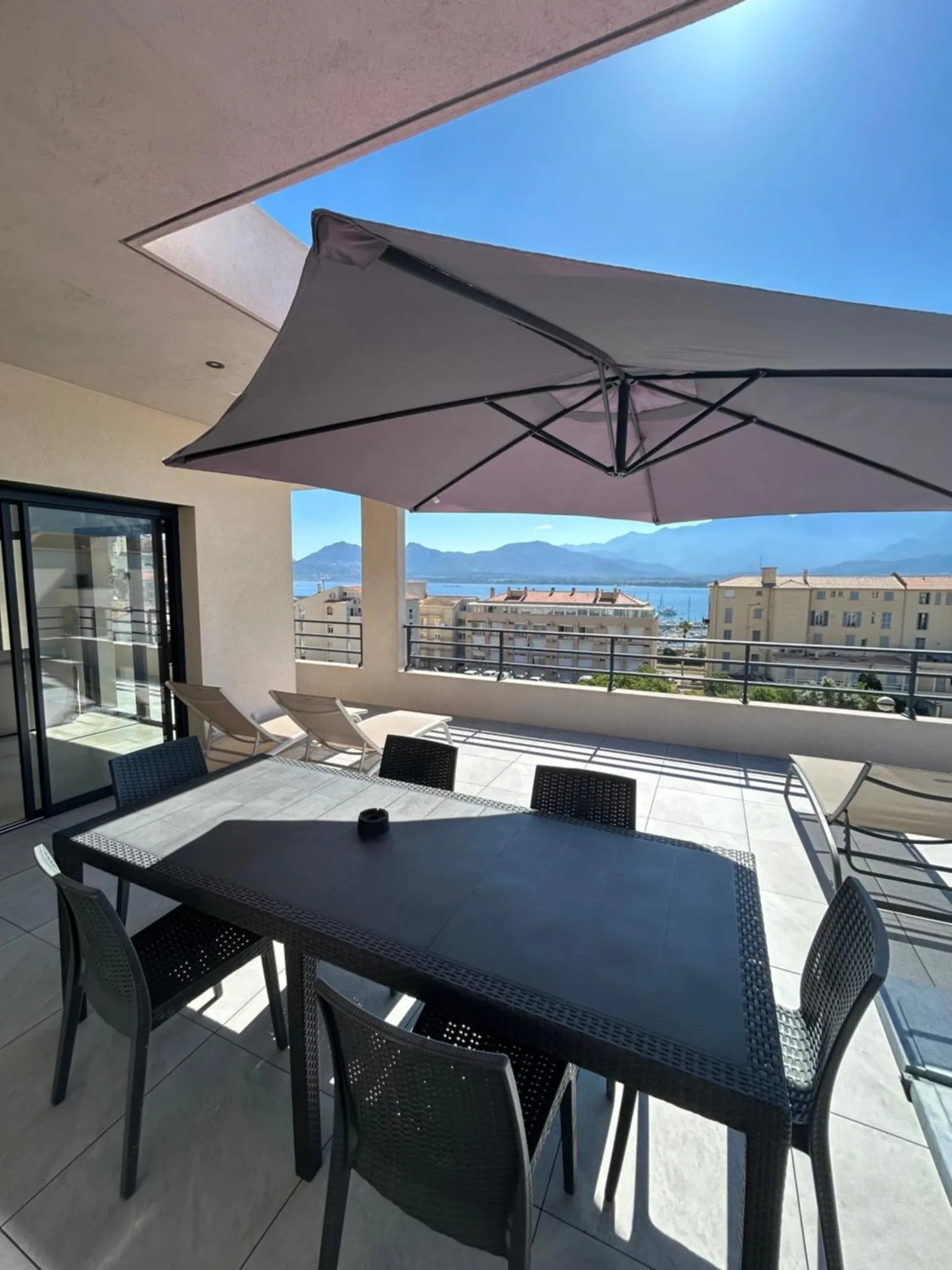 Une table et des chaises sont disposées sur une terrasse ombragée avec vue sur la mer et les montagnes.