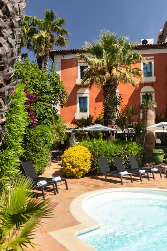 Transats et végétation luxuriante bordent une piscine ensoleillée devant un bâtiment aux tons chauds.