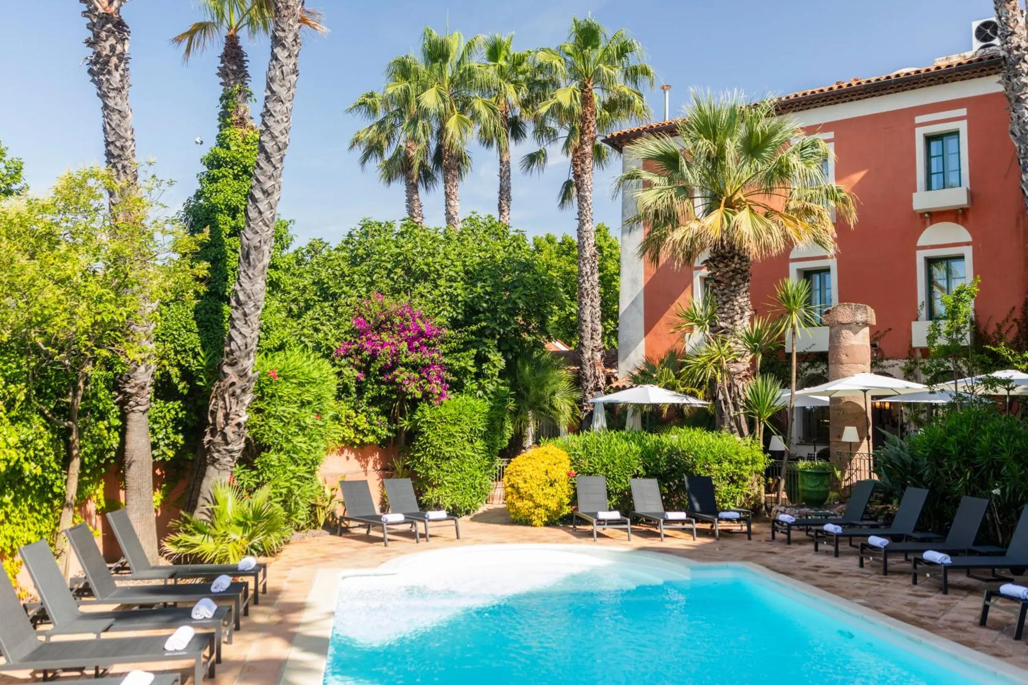 Transats et parasols bordent une piscine turquoise dans un jardin luxuriant.