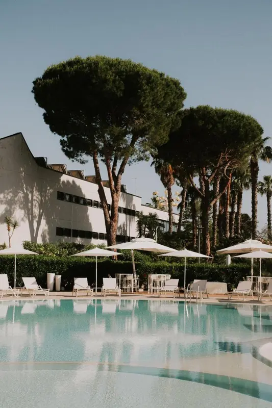 Transats blancs disposés autour d'une piscine turquoise sous des parasols, avec des arbres en arrière-plan.
