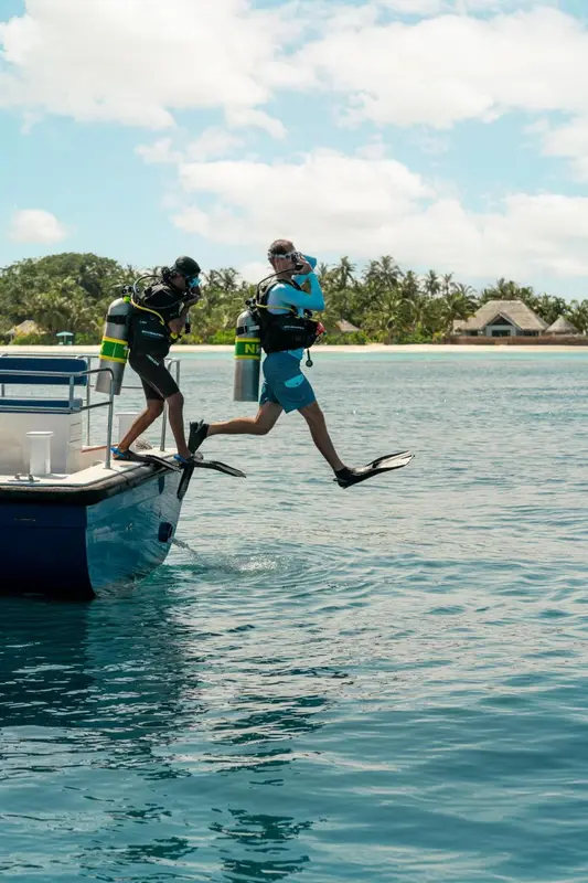 Deux personnes en combinaison de plongée s'apprêtent à sauter d'un bateau dans une eau turquoise.