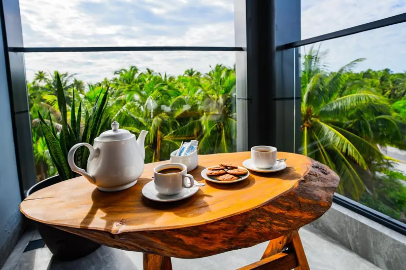 Une table en bois rustique avec une théière, des tasses de café et des biscuits, devant une grande fenêtre donnant sur une végétation luxuriante.