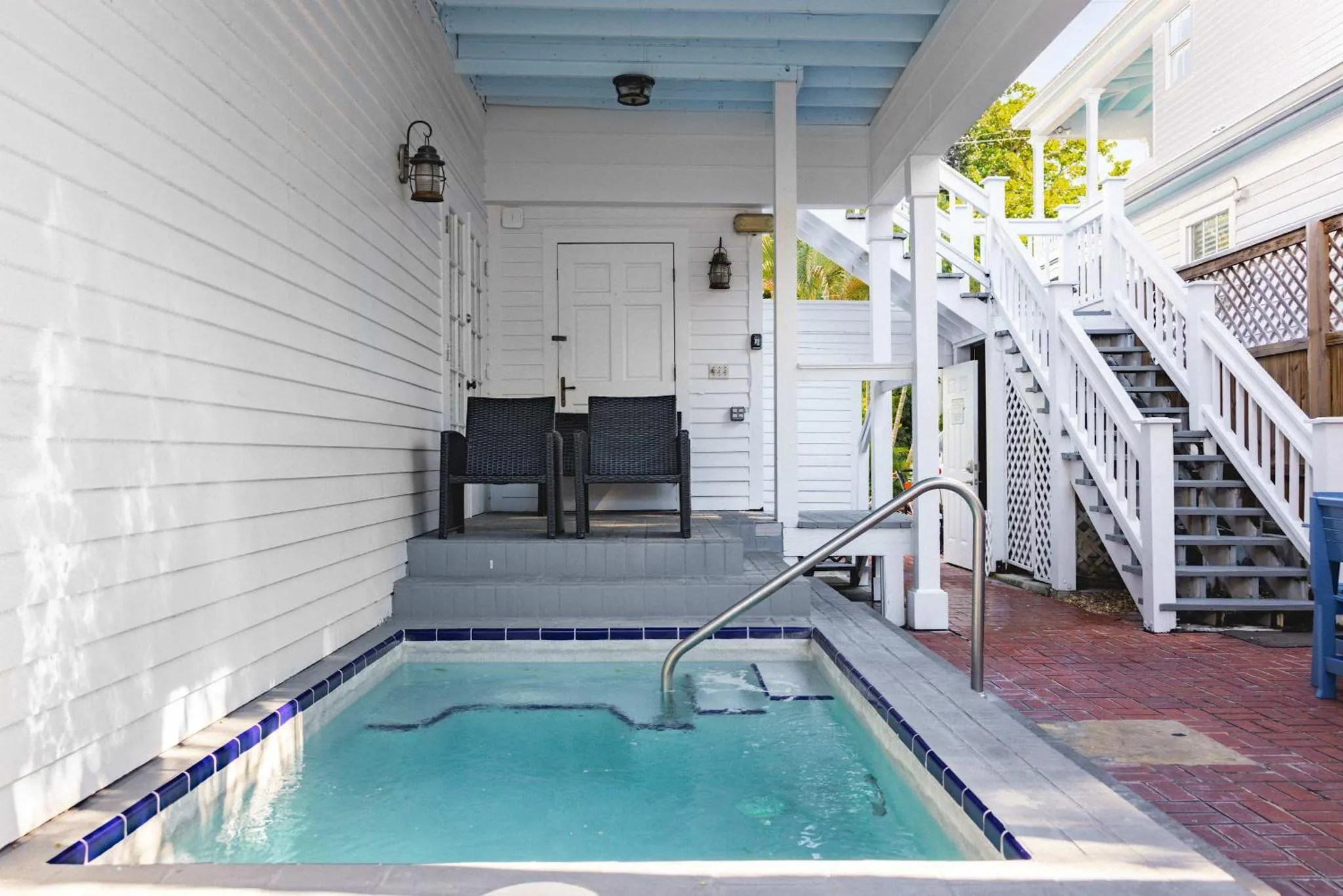 Petite piscine rectangulaire devant une maison blanche avec une terrasse et un escalier.